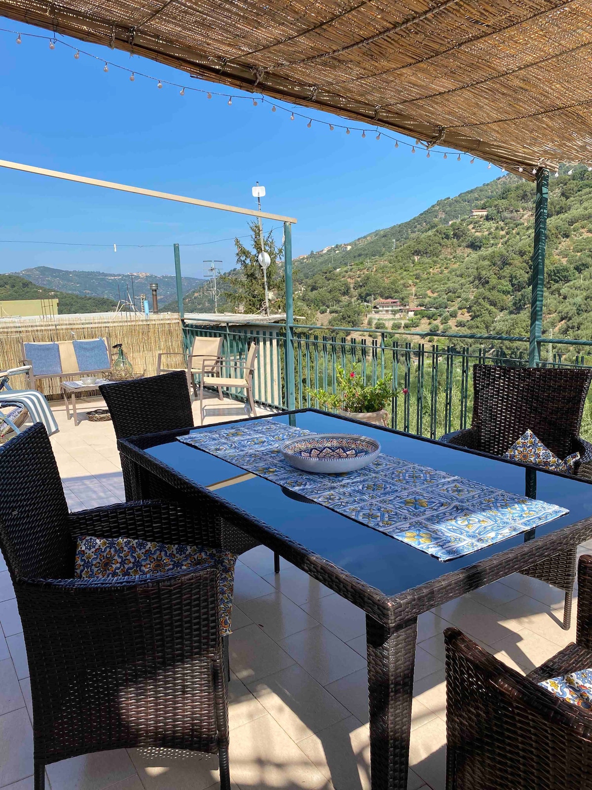 A spacious terrace is presented with a large dining table surrounded by four wicker chairs. A decorative table runner complements a central bowl. Sun loungers are visible to one side, with picturesque views of the surrounding hills in the background.