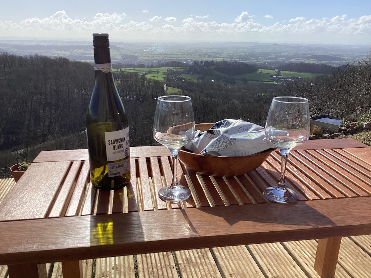 A wooden table is set with two glasses and a bottle of Sauvignon Blanc, overlooking a panoramic view of green fields and rolling hills. A bowl containing assorted snacks is also visible, enhancing the outdoor dining experience in a serene countryside setting.