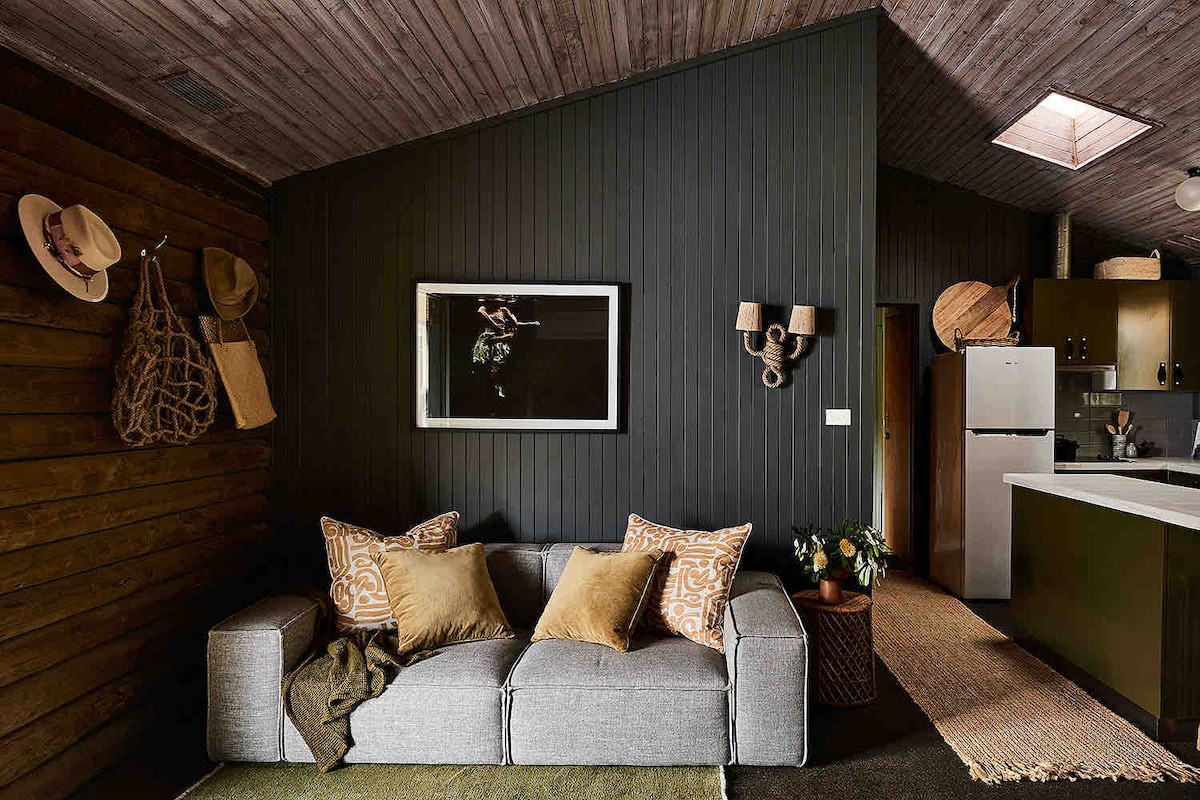 A cozy living area features a gray sofa adorned with patterned cushions, complemented by a textured throw. The wall is painted in a deep hue, and a framed artwork is displayed above. A small kitchenette is visible in the background, with a plant adding a touch of nature.