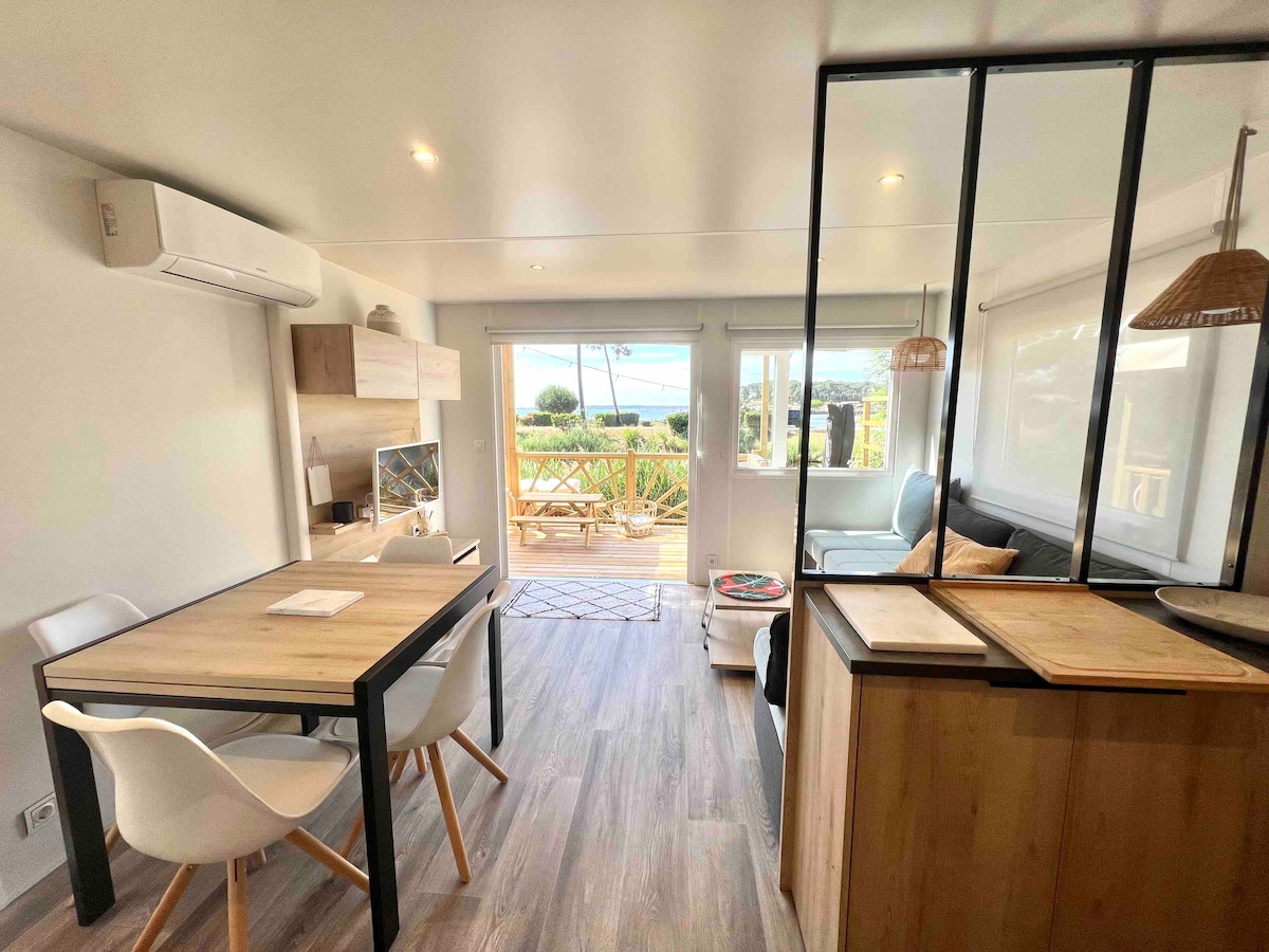 An open-concept living area features a dining table with white chairs, complemented by a wood and glass partition. Natural light illuminates the space through large sliding doors that open to a terrace, revealing views of greenery and distant water.