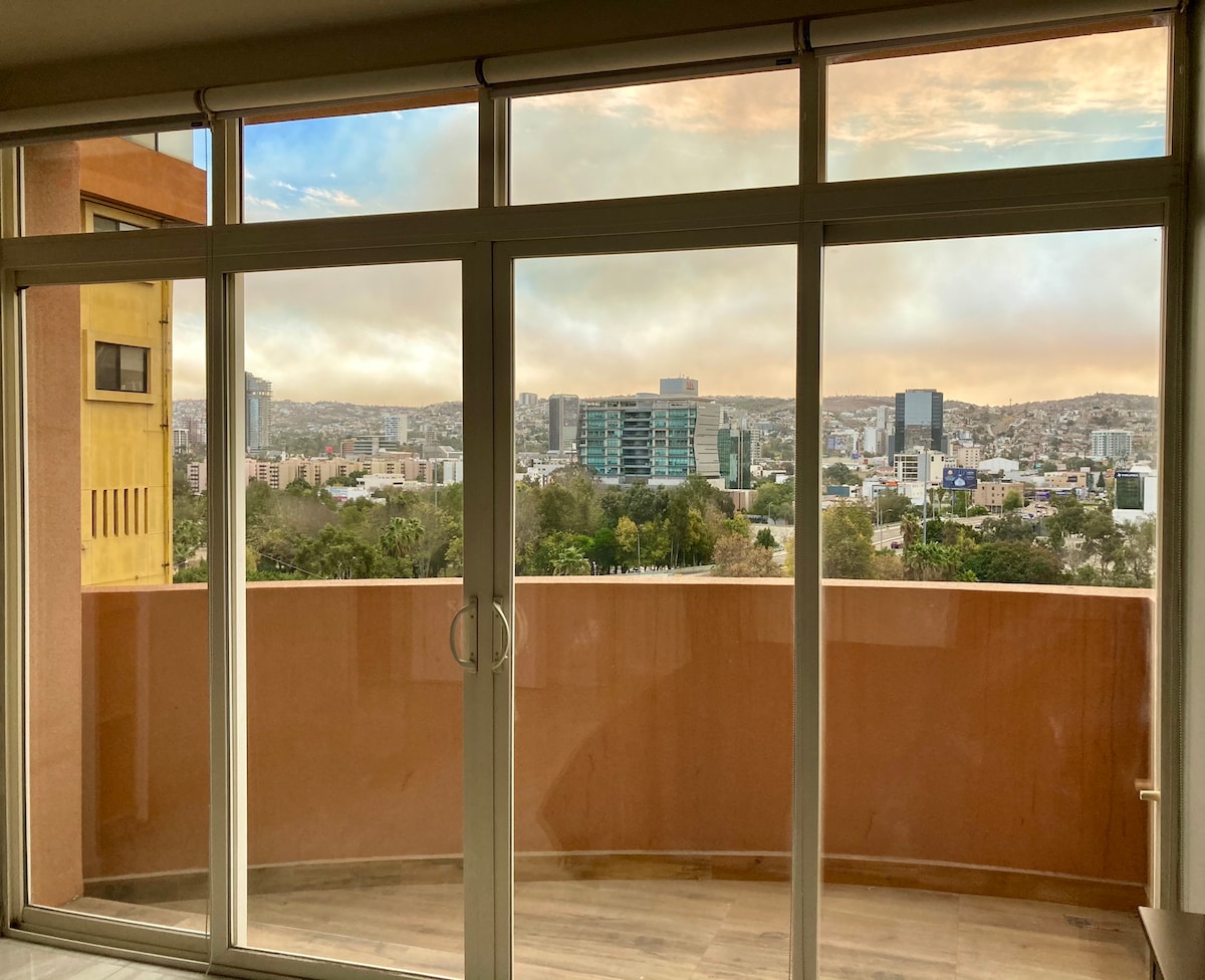 Large glass sliding doors reveal sweeping views of the cityscape, with buildings and hills extending towards the horizon. Soft natural light filters through the glass, while the balcony invites outdoor enjoyment in a quiet, modern environment.