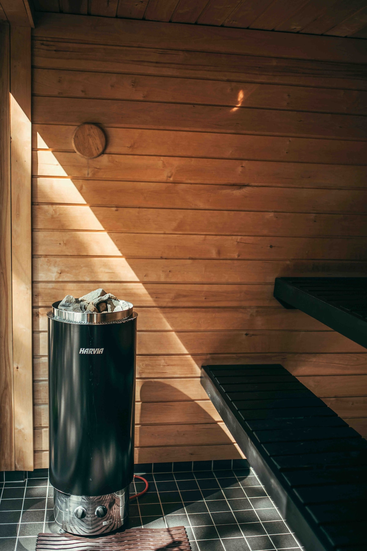 A modern sauna with wooden paneling creates a warm ambiance. A sleek black heater, topped with stones, stands beside a long wooden bench, offering ample seating. Soft sunlight filters through the space, accentuating the natural materials used throughout.