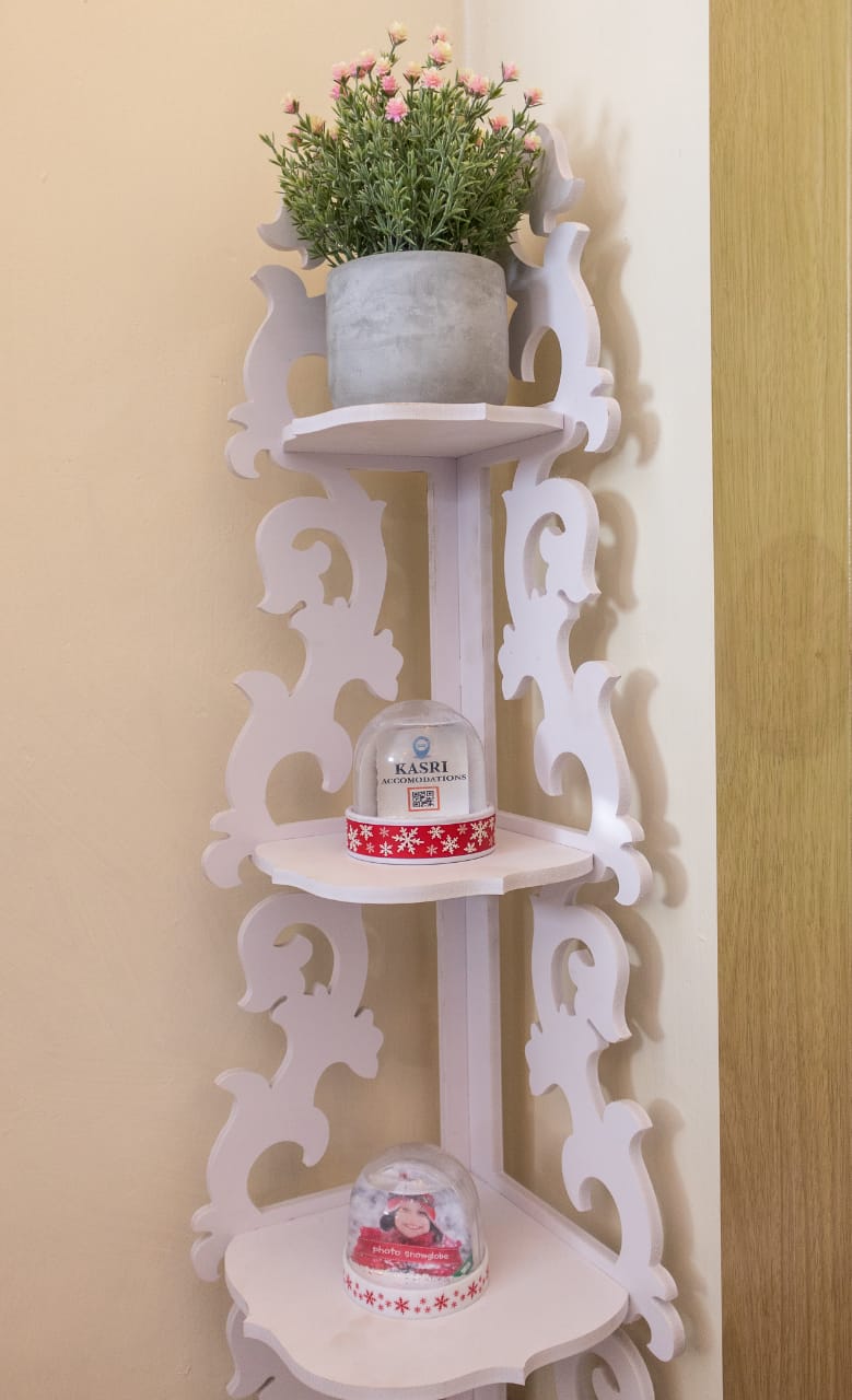 A decorative white shelving unit features three tiers, topped with a potted plant. A small snow globe is displayed on the second shelf, with another globe featuring a festive design on the bottom shelf. The background is a soft beige, enhancing the shelf's brightness.
