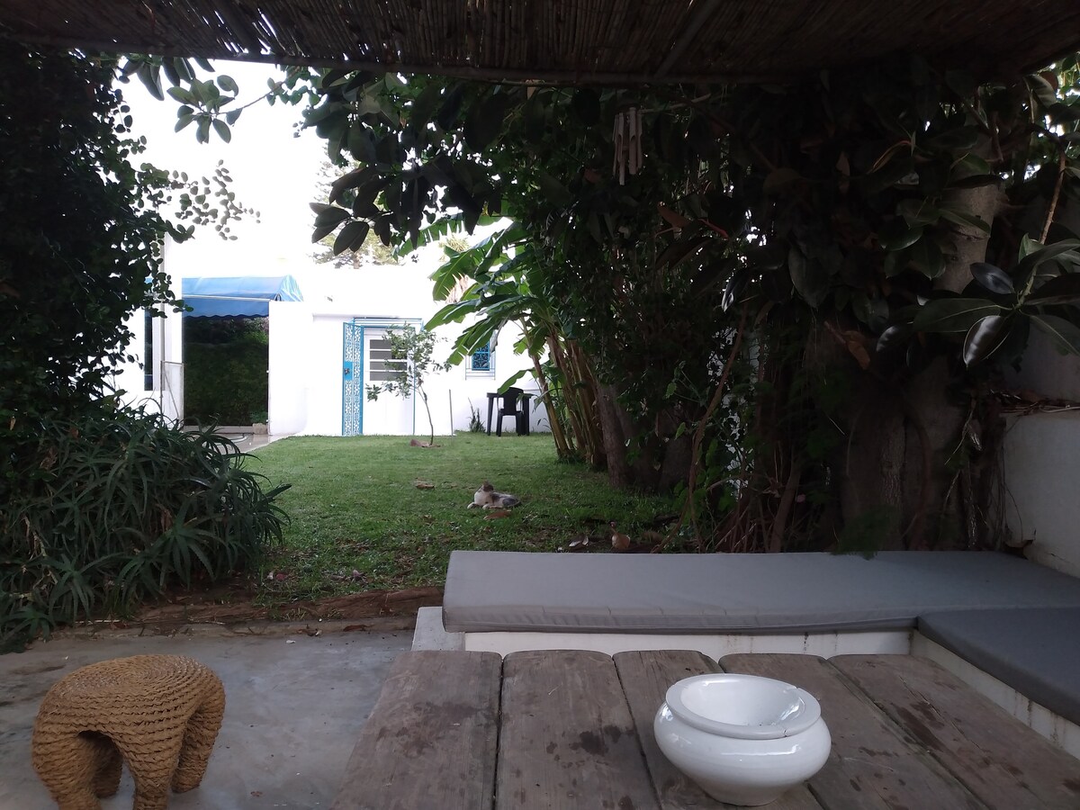 A serene outdoor space is framed by lush greenery, offering a well-maintained garden area. A wooden table and a woven stool are positioned in the foreground, while a shaded seating area is visible. In the background, a building is accessible through a pathway of grass.