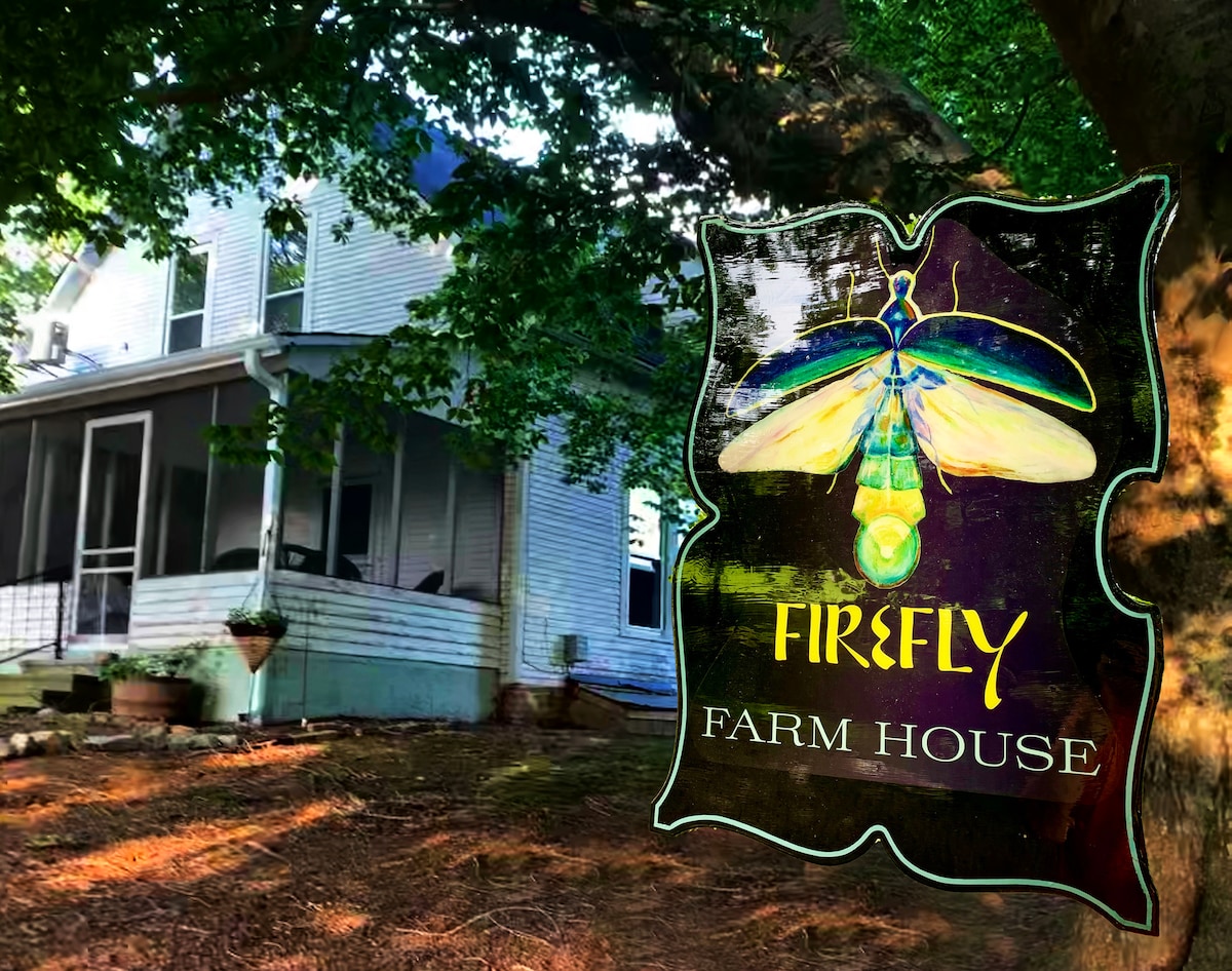 The exterior of Firefly Farmhouse is visible, showcasing a two-story structure framed by lush greenery. A large tree extends overhead, providing shade to the inviting porch. An artistic logo featuring a colorful firefly is presented in the foreground, enhancing the rural charm of the property.