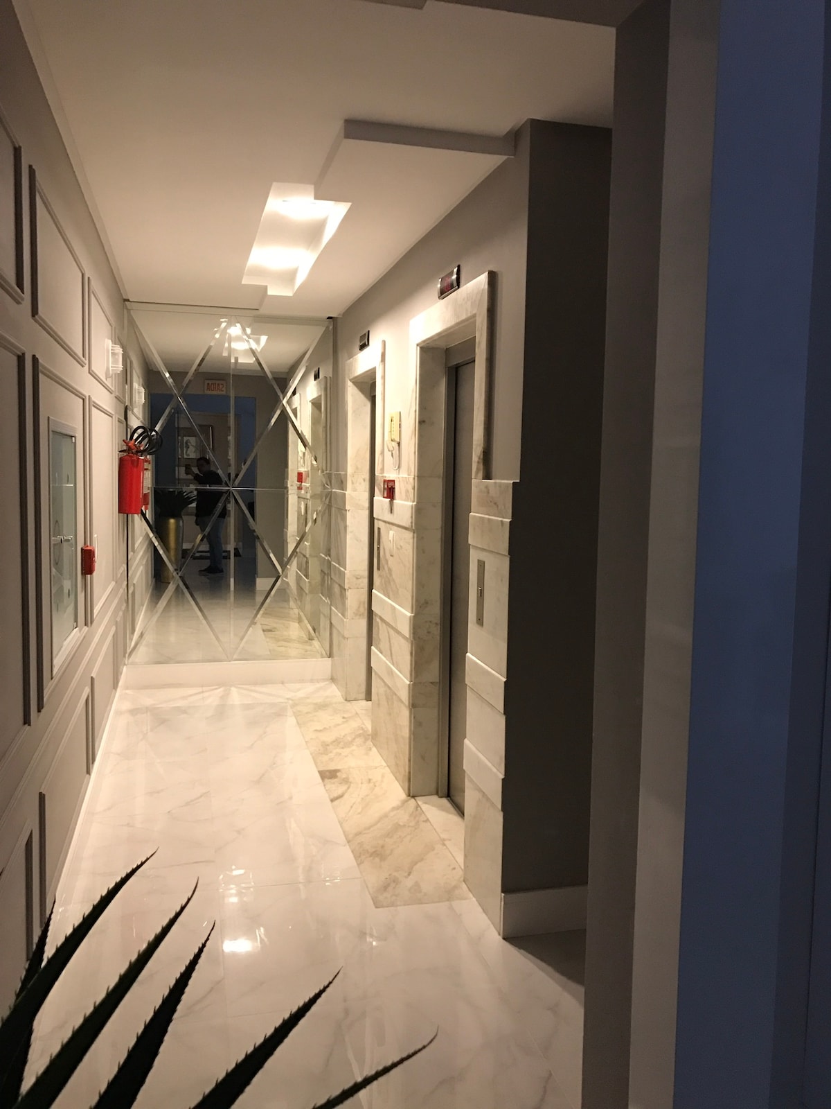 A well-lit hallway features glossy marble flooring and gray panelled walls. Modern elevators are visible at the end, with a glass partition adding elegance. Emergency equipment is neatly mounted on the wall, ensuring safety while maintaining a sleek design.