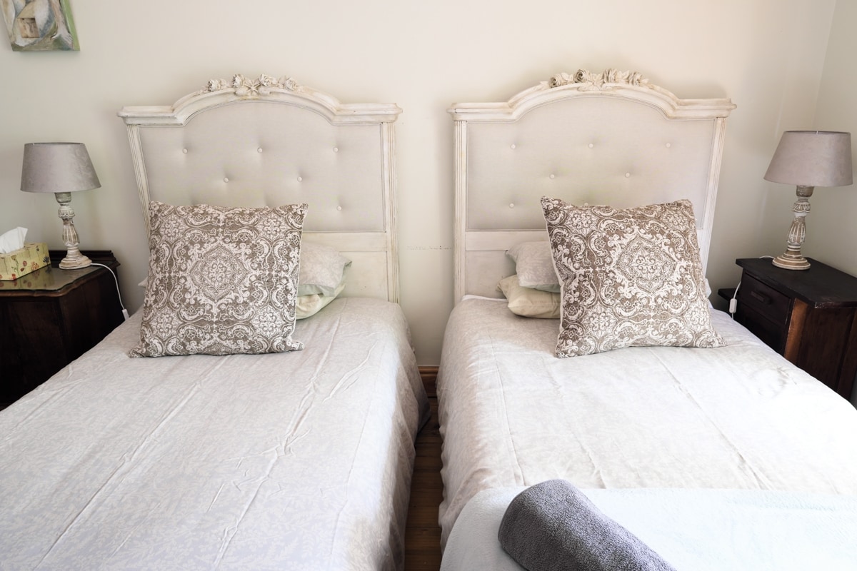 A room features two single beds, each adorned with decorative pillows and light-colored bedding. Bedside tables with lamps sit adjacent to the beds, providing both style and functionality in the cozy space.