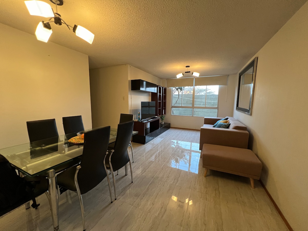 A spacious living area features a glass dining table surrounded by six dark chairs. A soft sofa sits beside a large window, offering ample natural light. A modern entertainment unit can be seen on one wall, enhancing the inviting ambiance of the room.