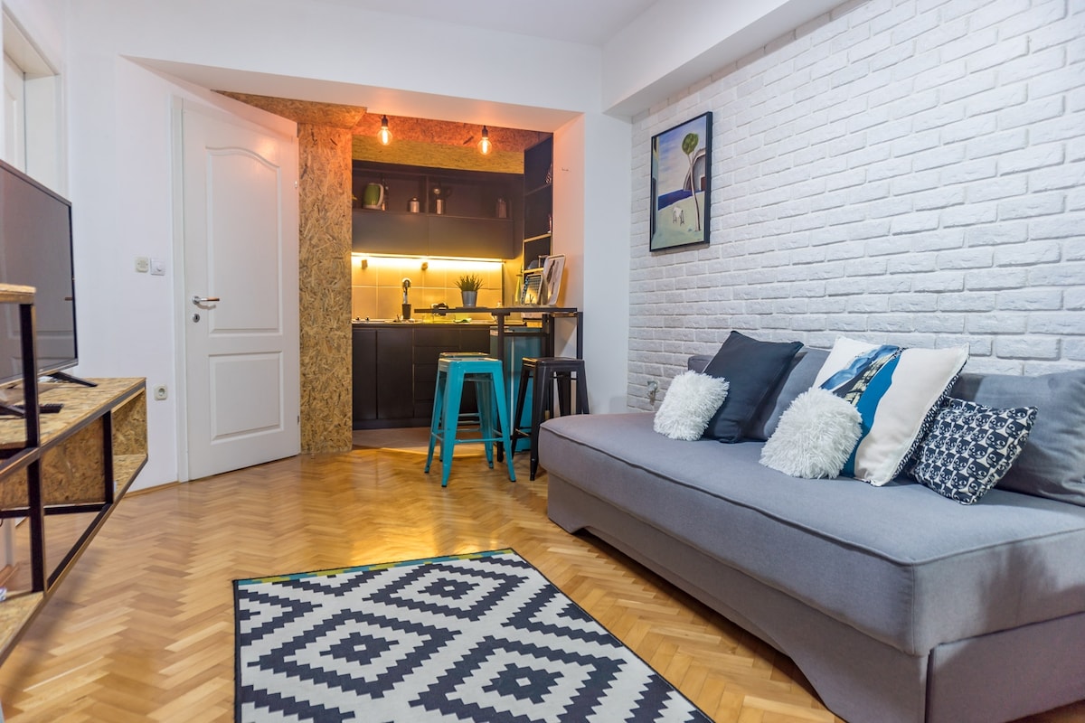 A cozy living area is presented with a spacious gray sofa adorned with decorative pillows. A patterned rug lies beneath, while a modern kitchenette is visible in the background. Soft lighting highlights the dark cabinetry, creating a welcoming atmosphere in this stylish space.