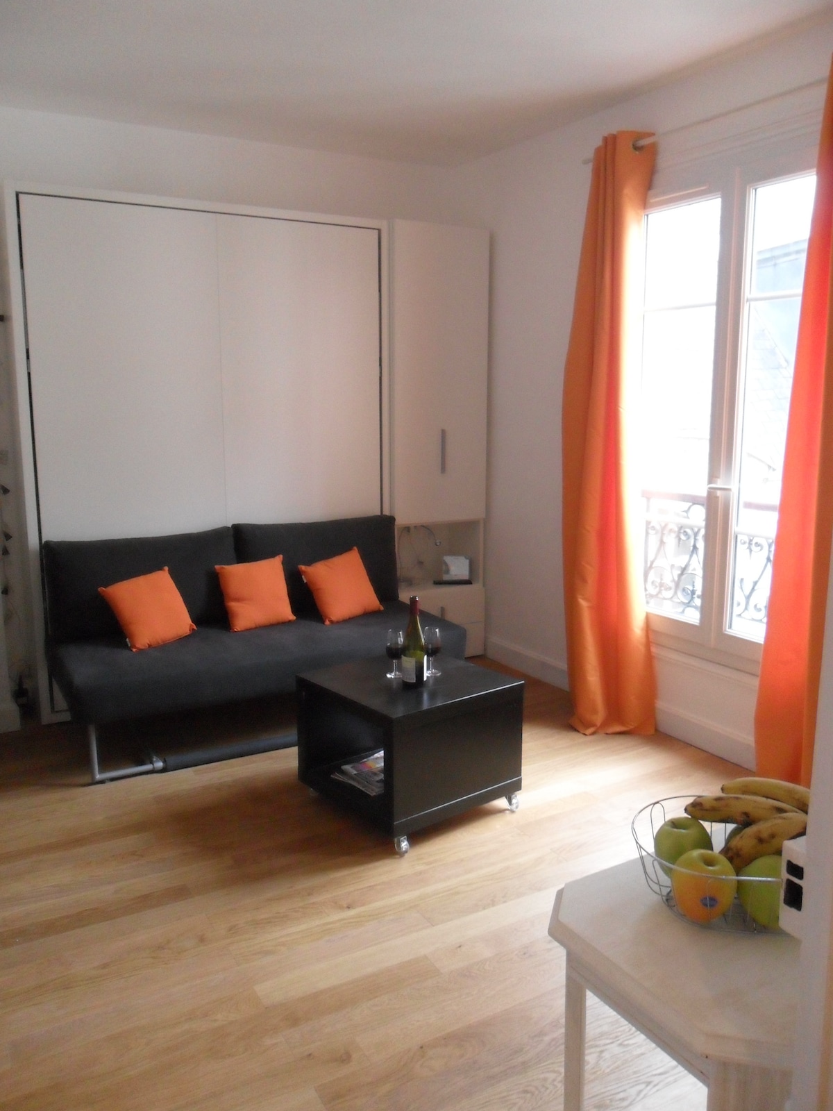 A compact living area is shown, featuring a gray sofa adorned with orange cushions. A dark coffee table sits in front, displaying a bottle of wine and two glasses. Natural light enters through a large window, complemented by soft orange curtains.