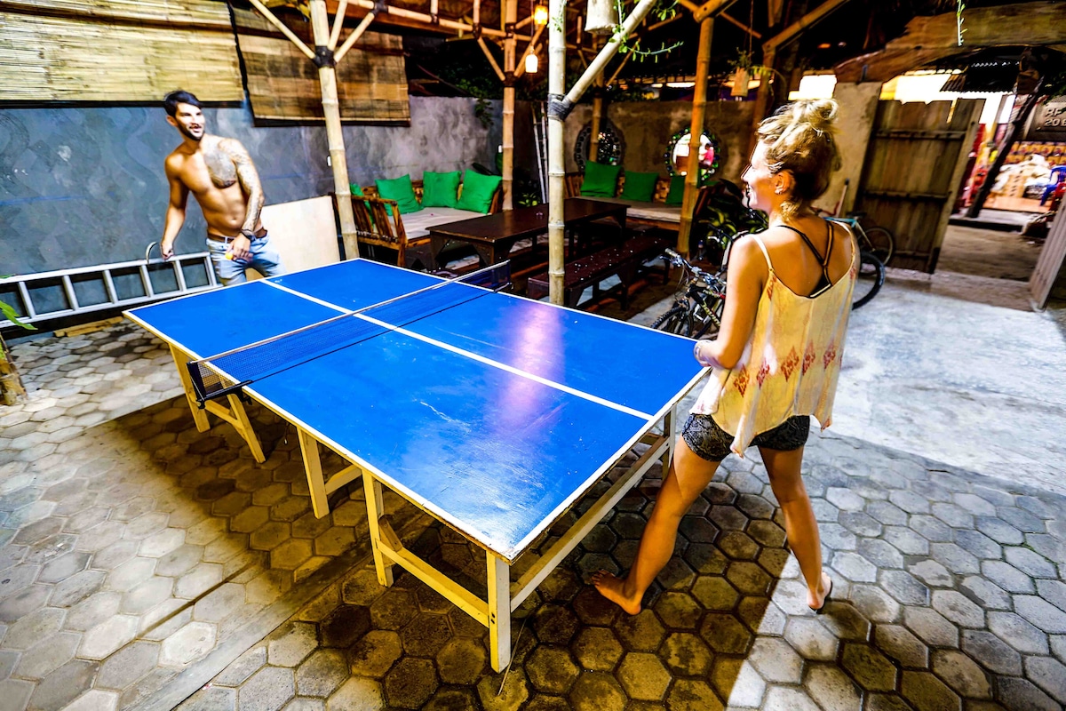 A vibrant communal area features a blue ping pong table in the center. Surrounding the table, rustic wooden furniture adds to the lively atmosphere. Open-air spaces are visible, with bamboo structures and greenery enhancing the social environment.