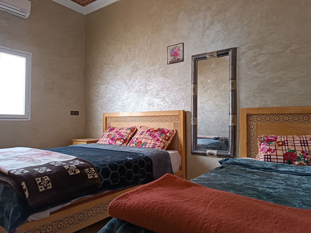 A well-appointed bedroom features two spacious beds, each adorned with patterned pillows and blankets. A decorative mirror is positioned against a neutral wall, while natural light enters through a nearby window.