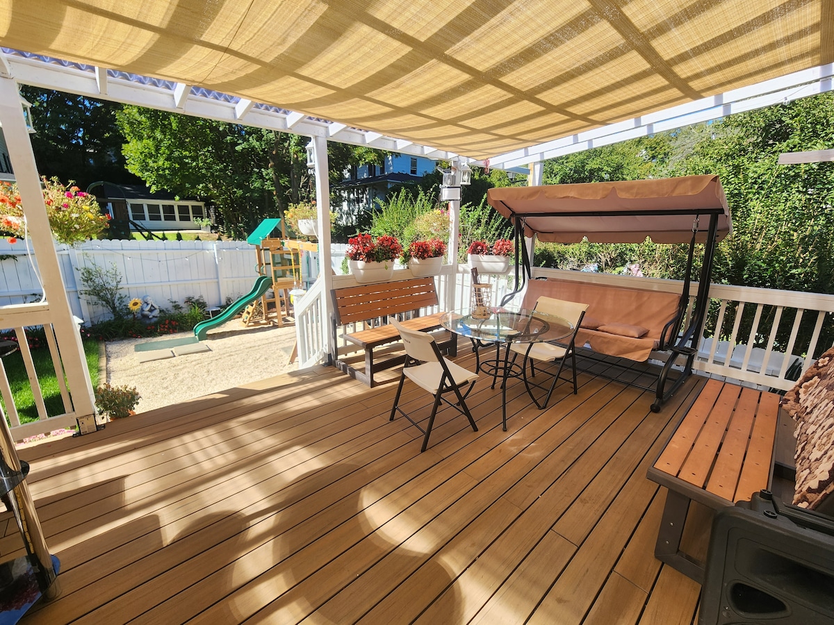 A spacious rear deck features a shaded seating area with a swing and dining set. Surrounding planter boxes are filled with colorful flowers. A playground area is visible in the background, enhancing the outdoor space.