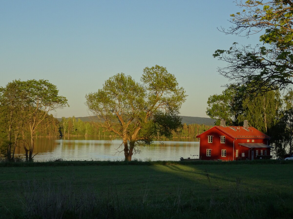 A red two-story house is situated near a serene lake. Large trees frame the scene, casting soft shadows on the grassy area. The tranquil water reflects the surrounding greenery, creating a peaceful atmosphere in the early evening light.