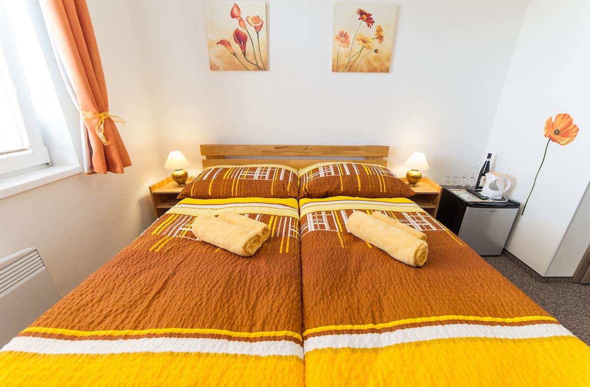 A double room features a double bed dressed in a vibrant brown and yellow quilt. Two matching towels are neatly placed on each side. Soft lighting from bedside lamps complements floral artwork on the walls. A minibar sits discreetly within the room's layout.