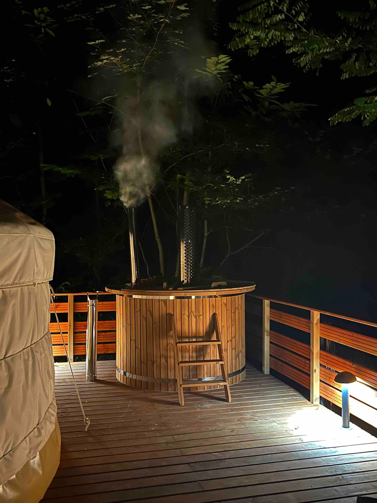 A wooden hot tub sends steam into the cool night air, surrounded by greenery. The outdoor terrace is illuminated by soft lighting, allowing for a peaceful ambiance. A wooden chair is positioned nearby, enhancing the serene environment for relaxation.