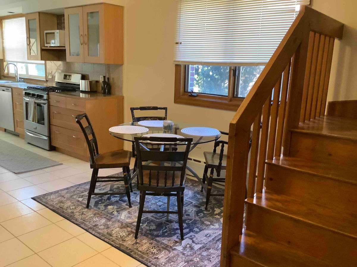 A dining area is showcased, featuring a round table surrounded by four black wooden chairs. A patterned rug lies beneath the table, and a window allows natural light to filter in. A staircase is visible in the background, leading to the upper level.