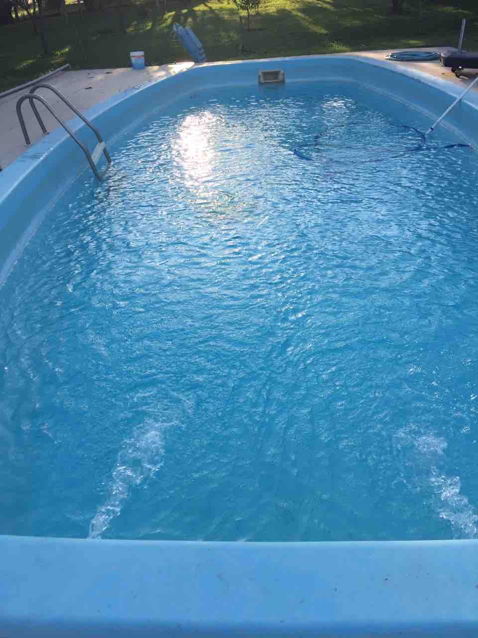An outdoor swimming pool is depicted, featuring clear, blue water reflecting sunlight. The pool edges are lined with light blue tiles, and a set of stainless-steel steps leads into the water. Surrounding greenery provides a serene setting.