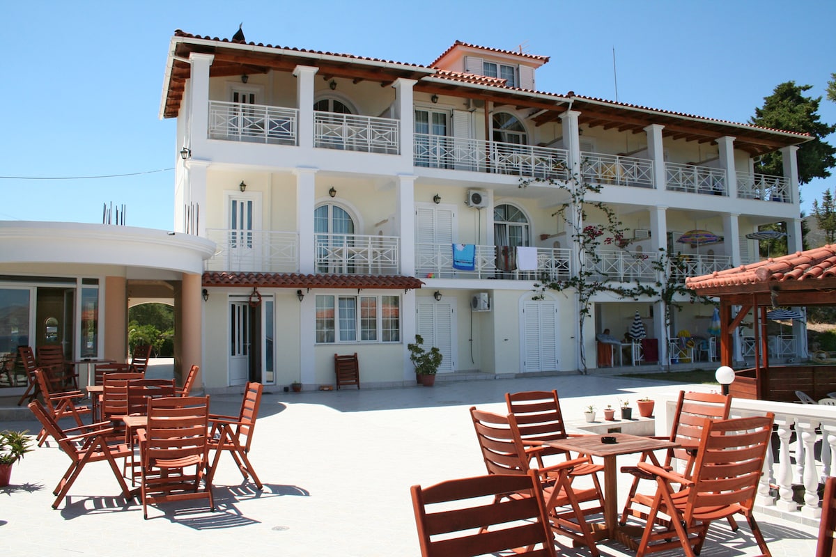 A three-story building is presented with a white exterior and a terracotta roof. Balconies feature decorative railings, and a spacious patio with wooden furniture is visible in the foreground, providing a welcoming space for relaxation.