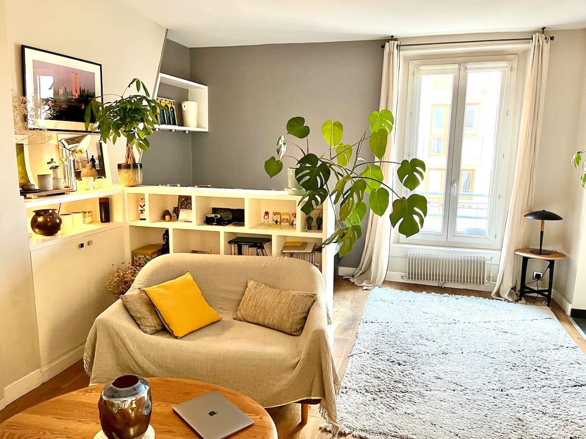 A bright and calming living area is filled with natural light from a large window. A cozy couch, adorned with a yellow cushion, is paired with a wooden coffee table. Green plants add a touch of nature, while a light rug warms the wooden floor.