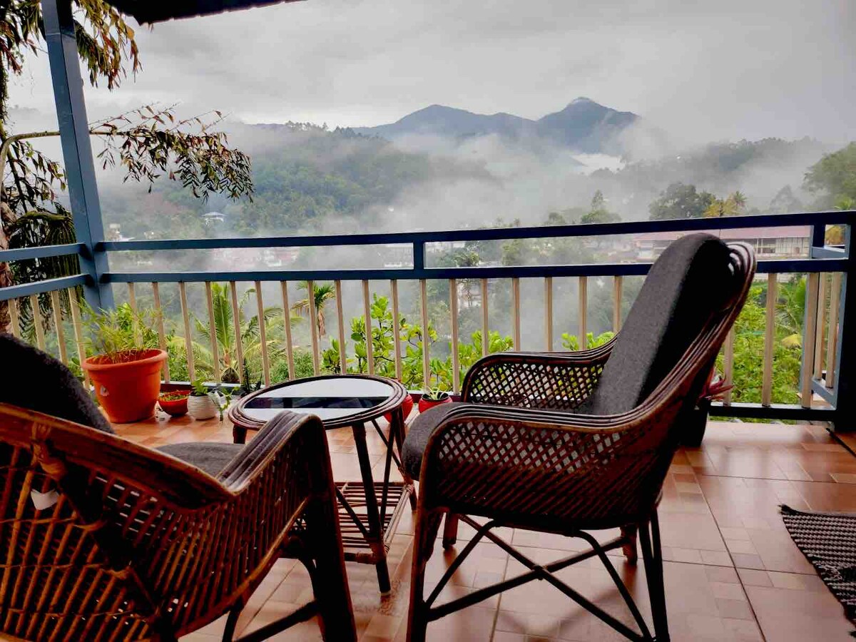 A cozy balcony is equipped with two woven chairs and a glass table, offering views of mist-covered mountains and lush greenery in the distance. Potted plants are placed elegantly on the railing, adding a natural touch to the tranquil setting.