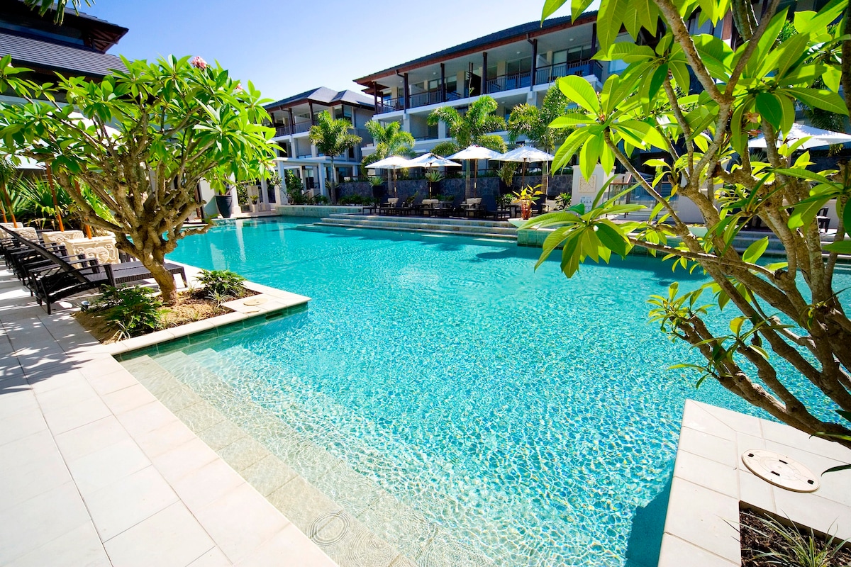 A spacious tropical pool is framed by lush greenery and sun loungers, with umbrellas providing shade. The crystal-clear water reflects the bright blue sky, inviting guests to relax by the poolside. The modern architecture of the building is visible in the background.