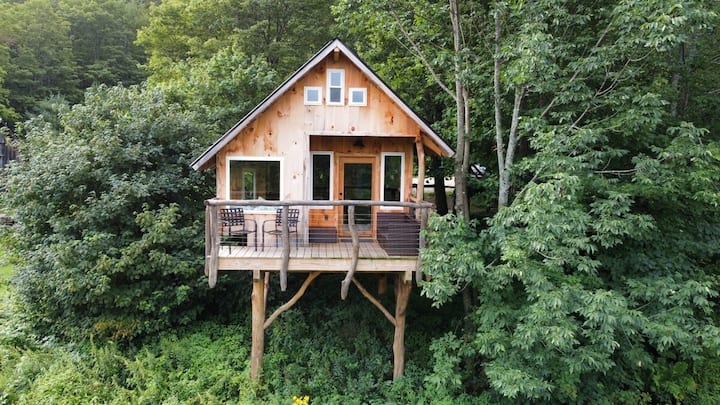 A Vermont treehouse sits high above the ground and nestled in the tree canopy with a lovely patio in front with outdoor seating.