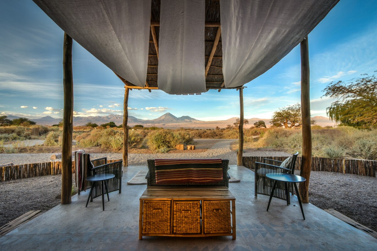 An outdoor seating area features a wooden coffee table and two chairs, surrounded by natural landscapes. A canopy covers the space, offering shade while providing views of distant mountains under a soft sky. The surrounding area consists of desert vegetation.