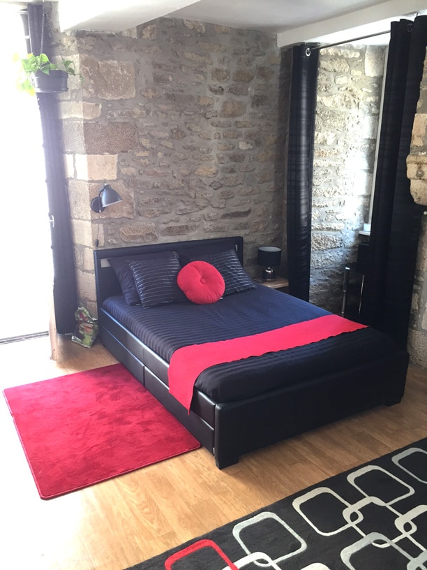 A spacious sleeping area features a neatly made bed with dark bedding and a bright red throw blanket. A round red pillow adds a pop of colour. Thick black curtains frame a window, while a small lamp and a rug complete the cozy atmosphere.