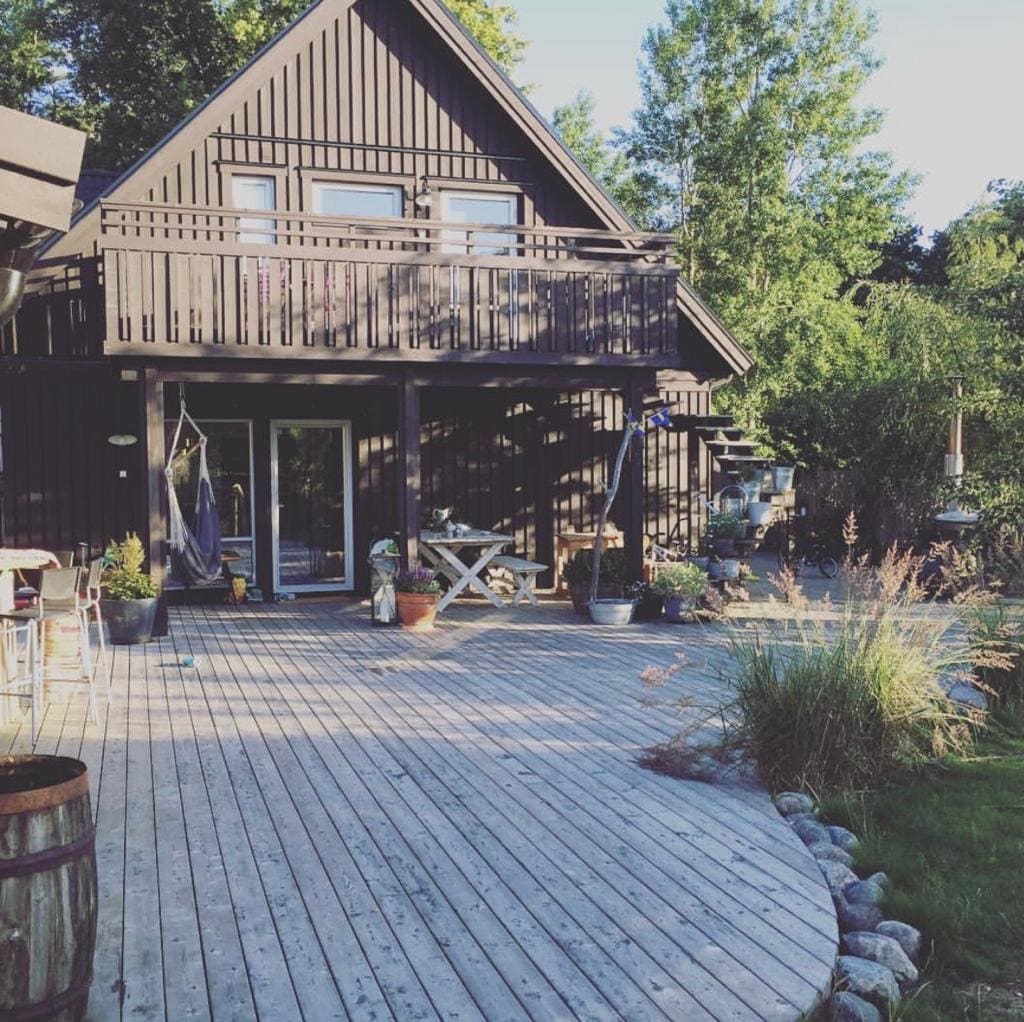 A spacious wooden deck surrounds a two-story dark wooden house, featuring large glass doors and a balcony. Surrounding greenery enhances the natural setting, while various potted plants add to the outdoor decor. Pathways lead through the garden, inviting exploration.