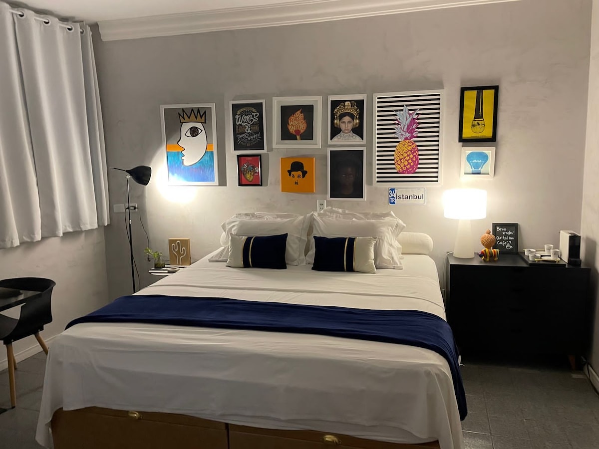 A comfortable bedroom is featured, showcasing a spacious bed with white linens and decorative pillows in black and blue. An array of framed artworks adorns the wall above the bed. A bedside table with a lamp and a black chair complete the cozy setup, with natural light filtering through white curtains.