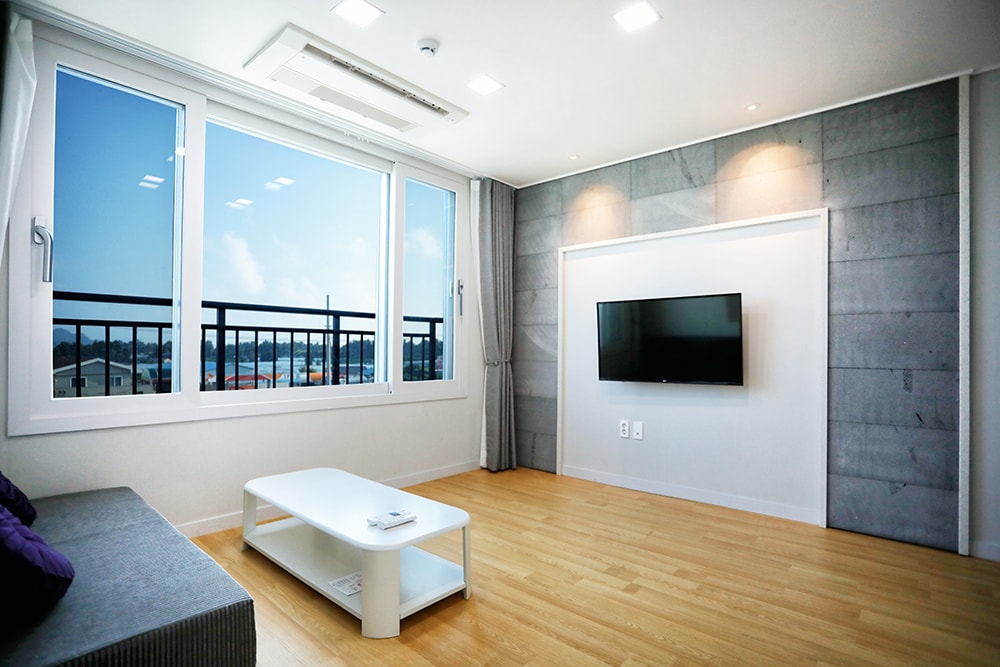 A light-filled living area features large windows offering scenic views. A flat-screen television is mounted on a smooth wall, complemented by a low coffee table positioned on warm wooden flooring. A comfortable sofa in soft tones invites relaxation.