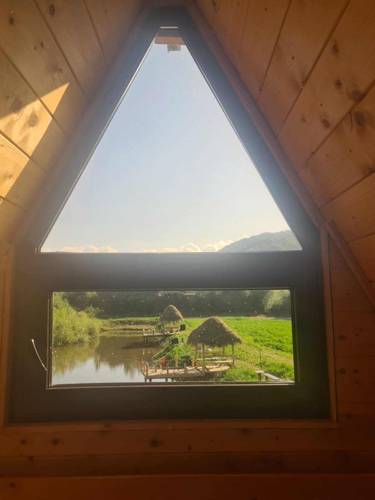 A large triangular window frames a scenic view of a serene lake and nearby dock. The vibrant green landscape is dotted with rustic huts, and distant mountains provide a tranquil backdrop against a clear sky.