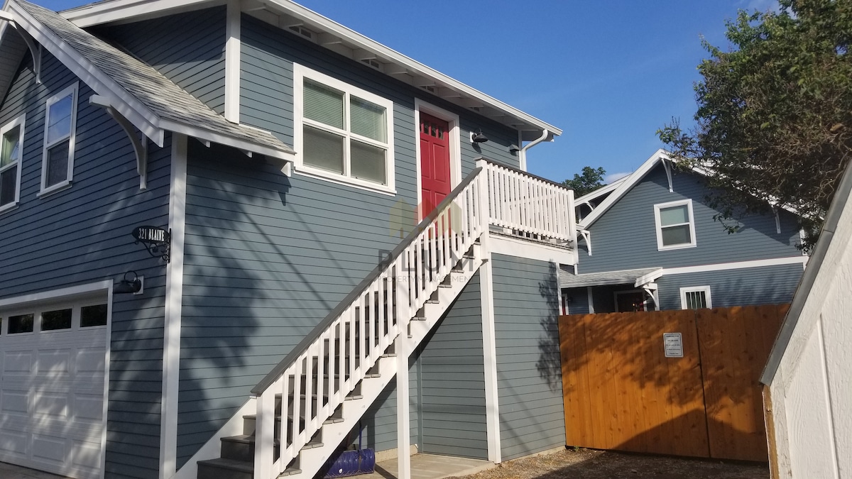 A two-story guest suite with a modern blue exterior is highlighted by a bright red door. A white wooden staircase leads to the entrance, while a fenced area is visible to the side, offering privacy. Clear skies and natural light enhance the welcoming appearance.