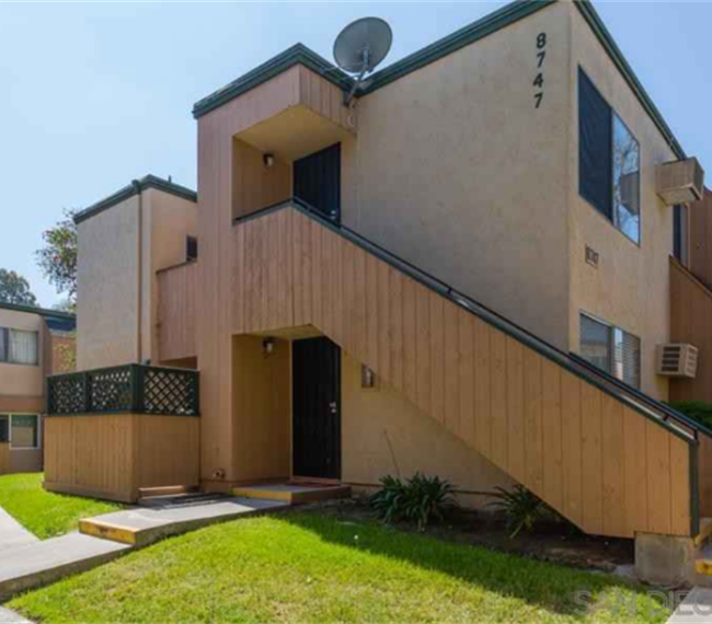 An exterior view of a two-story apartment building is presented. The entrance features a set of stairs leading to a door, flanked by large windows. Surrounding greenery adds a touch of nature, while a small patio area is visible beneath the stairs.