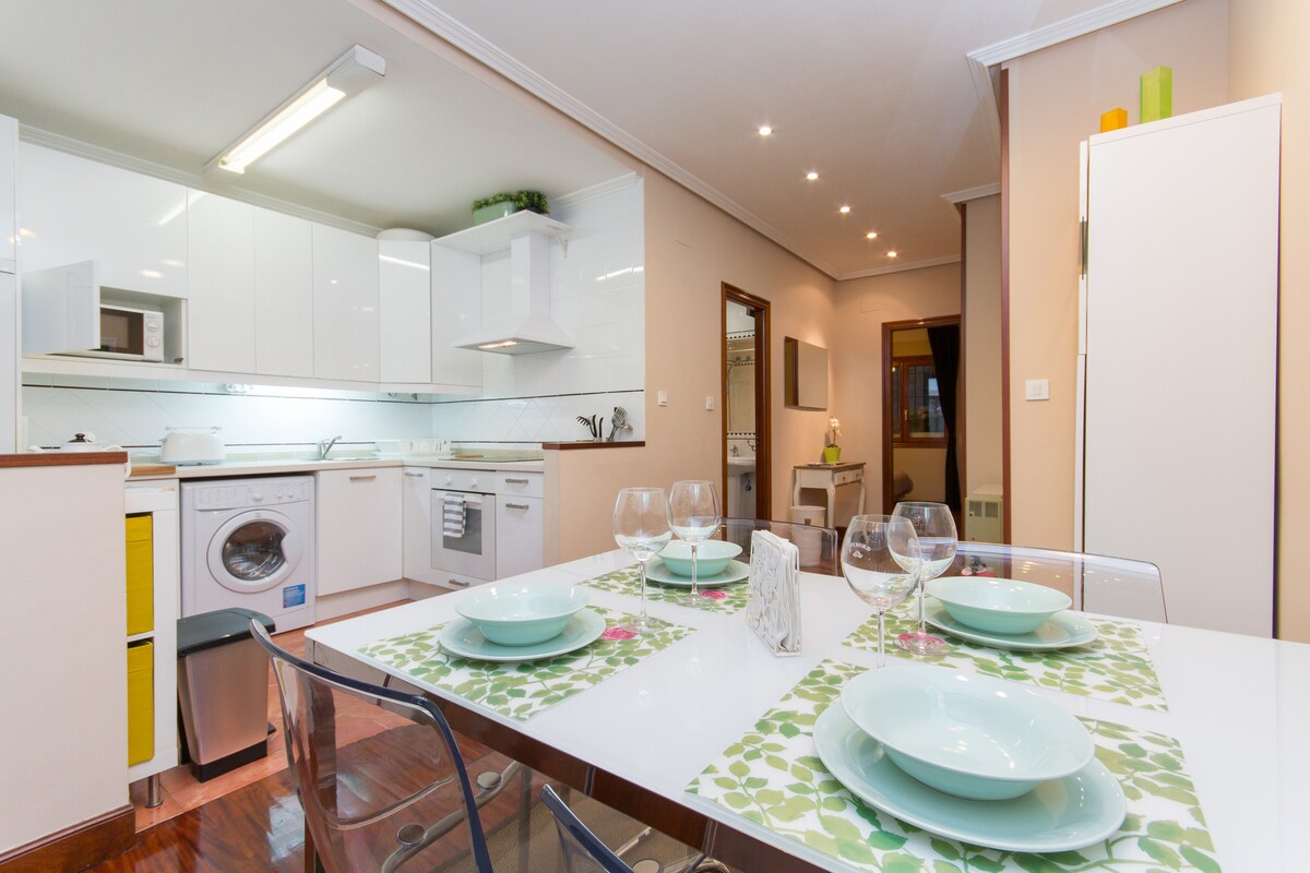 A modern kitchen and dining area are showcased, featuring a white cabinetry with sleek lines. A dining table is set with green-patterned placemats, clear dishware, and an array of table settings. Natural light illuminates the space, highlighting the contemporary design.