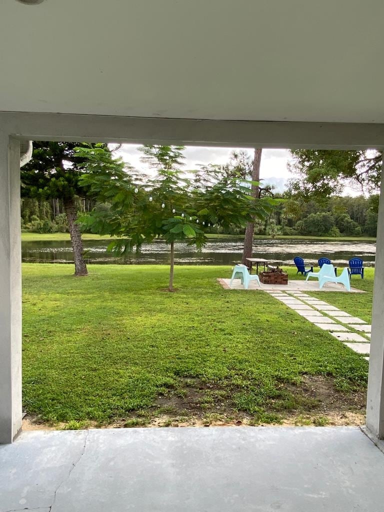A view of a serene lake surrounded by greenery is captured through an archway. A small tree stands nearby on a grassy area, with a stone pathway leading to colorful outdoor chairs. The scene presents a peaceful outdoor space for relaxation.