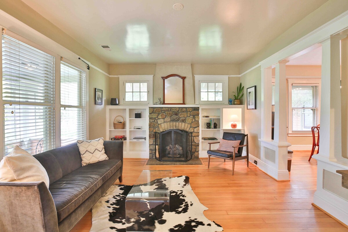 A spacious sitting room features a modern gray sofa positioned in front of a stone fireplace. Natural light enters through large windows with white blinds, illuminating the hardwood flooring. A unique cowhide rug lies on the floor, complementing a stylish armchair nearby.