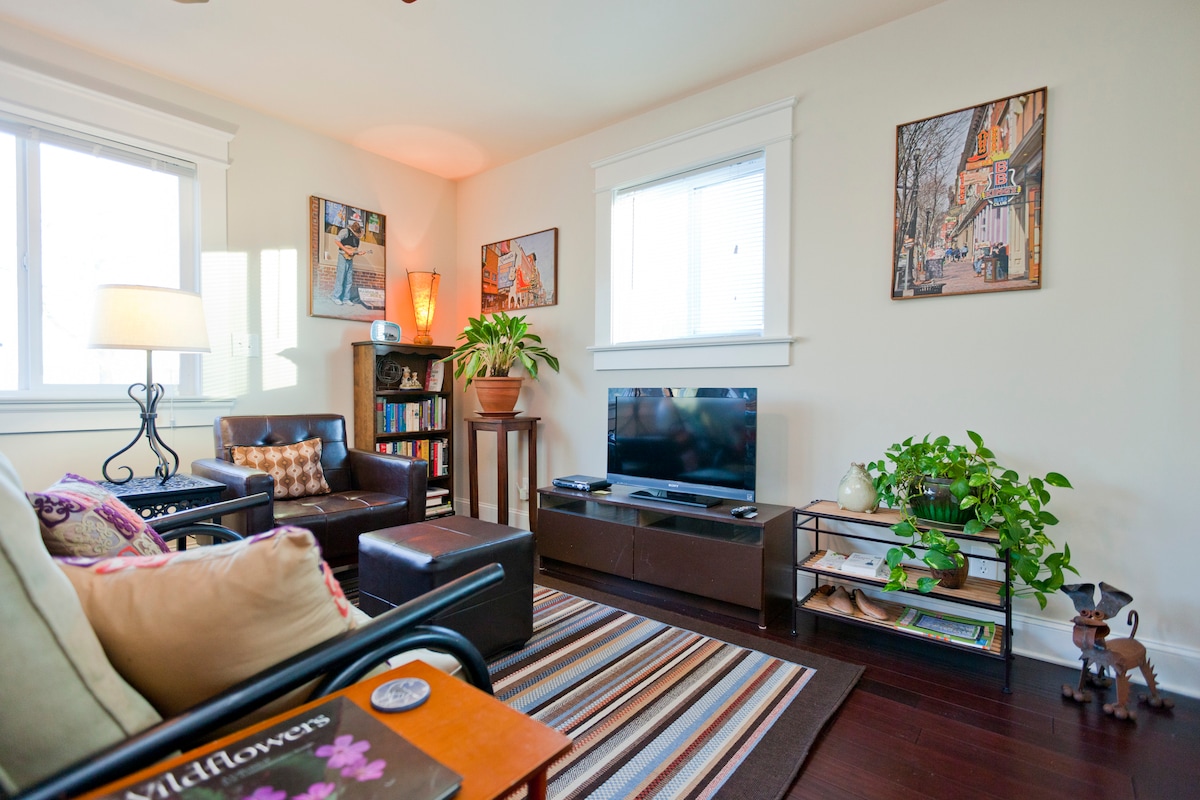 A bright living area is presented with a comfortable sofa and a leather armchair. Two framed artworks adorn the walls, while a small television is placed on a low media console. Natural light streams in through the window, and decorative plants add greenery.