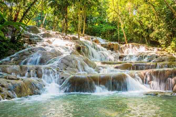 A cascading waterfall flows over a series of rocky tiers, surrounded by lush greenery. The water flows into a tranquil pool at the base, reflecting the sunlight filtering through the trees above.