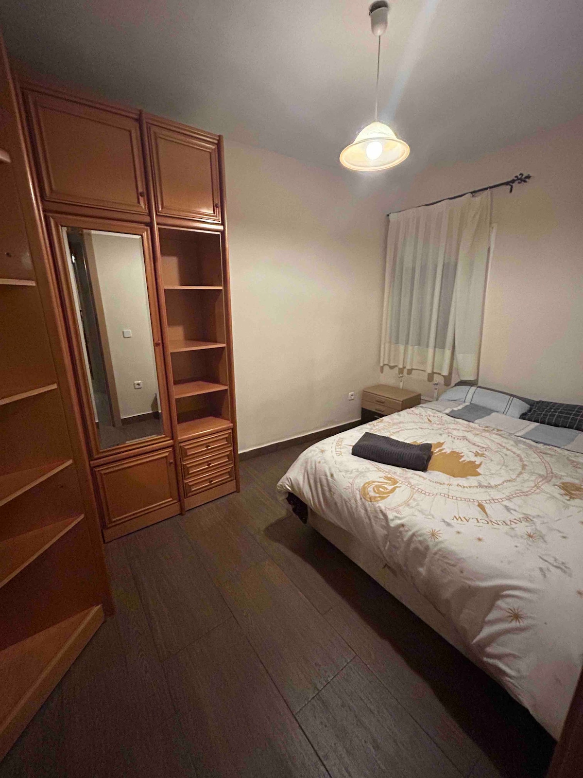 A cozy bedroom is presented with a spacious bed dressed in light bedding, accompanied by a folded towel. A wooden wardrobe with shelves and drawers stands along one wall, while light-colored curtains frame a window. Warm light is provided by a ceiling fixture.