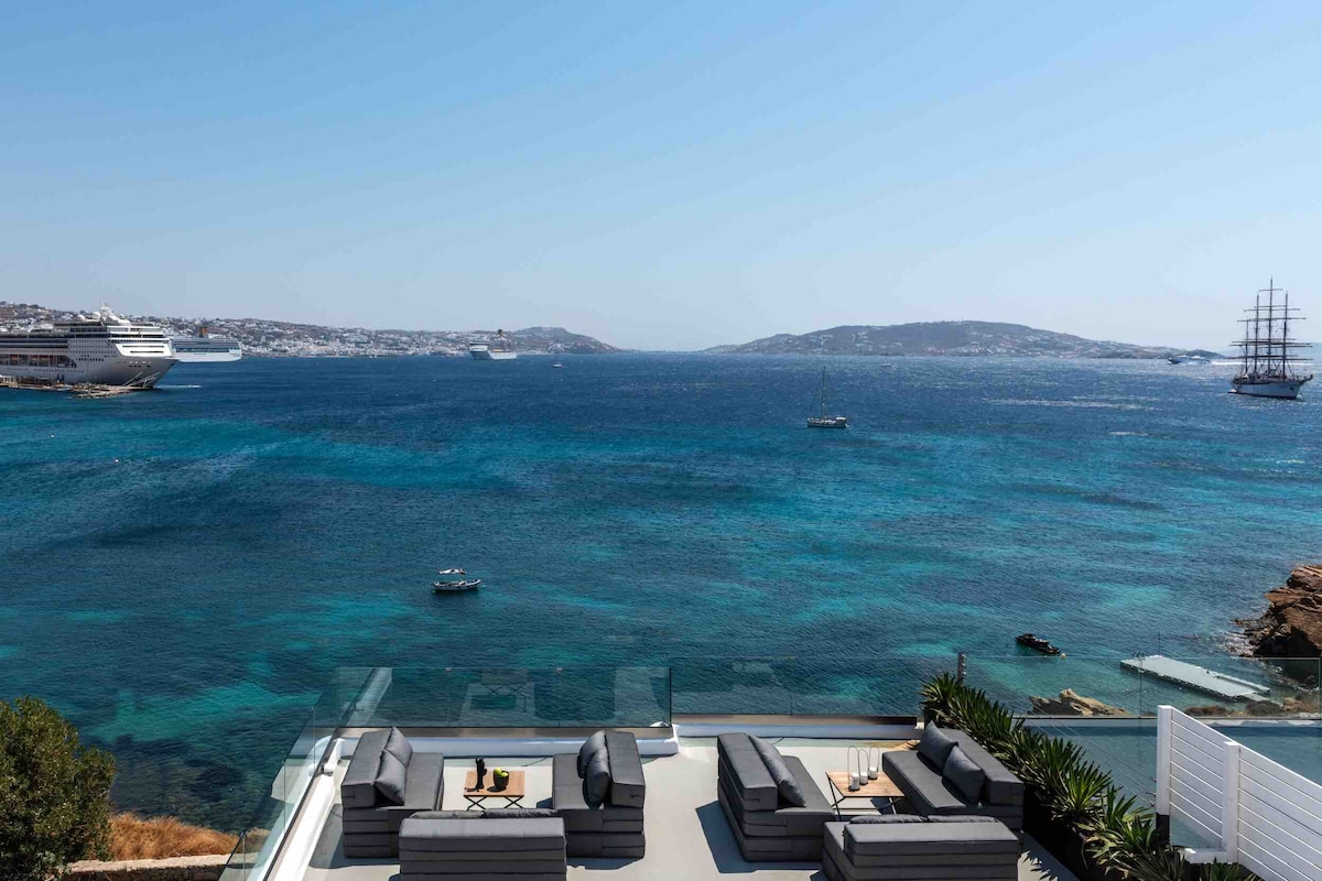 A spacious terrace offers an unobstructed view of the Aegean Sea, with various shades of blue blending seamlessly. Comfortable outdoor seating arrangements invite relaxation, while boats are seen navigating the water in the distance. The horizon features the skyline of Mykonos town under clear skies.