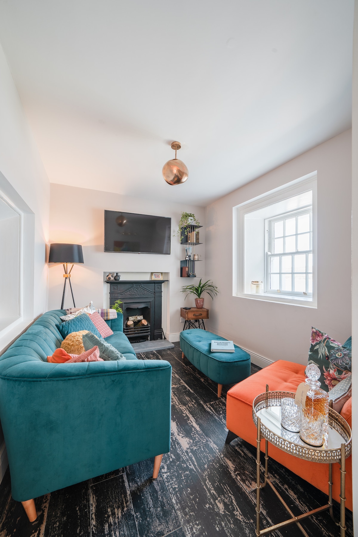 A cozy living area features a blue-green velvet sofa and an orange accent chair, complemented by a small coffee table with decorative items. A flat-screen TV is mounted above an elegant fireplace, while a window provides natural light, and shelves display plants and decor.
