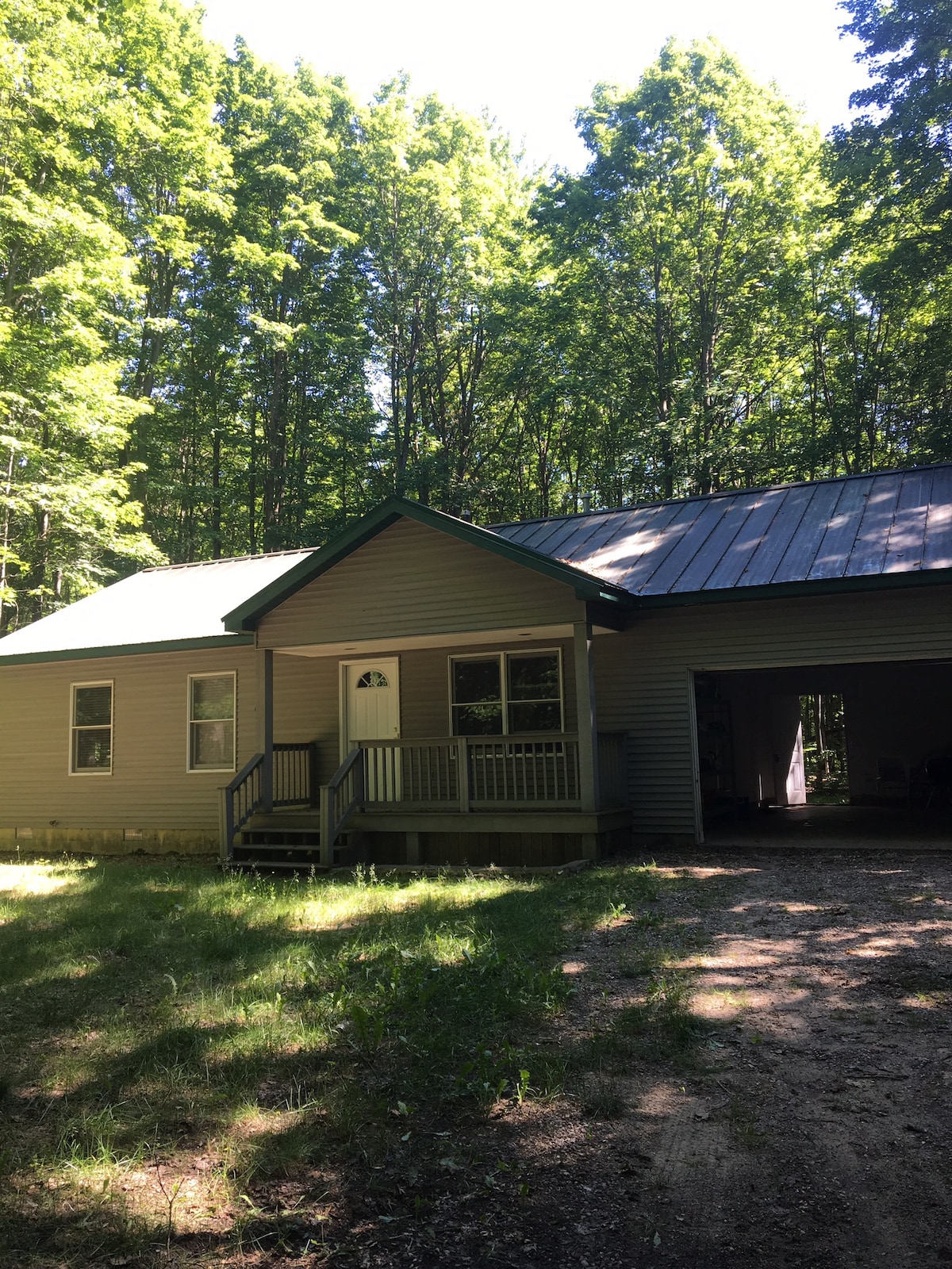 A single-story house is nestled among tall trees, featuring a covered front porch with steps leading to the door. The structure boasts a metal roof and a spacious garage to the side, surrounded by greenery and a natural landscape.