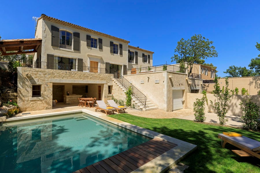 The exterior of the property showcases a well-maintained garden and a heated swimming pool. The house has a traditional architectural style with a light-colored façade and multiple balconies. Sun loungers are placed near the pool, and the area is surrounded by greenery and stone pathways.