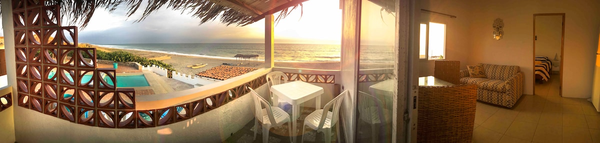 A panoramic view showcases the beach and ocean at sunset from a private patio. A dining table with chairs is positioned on the patio, offering an inviting space. The interior features a comfortable seating area, with light filtering through large glass doors.
