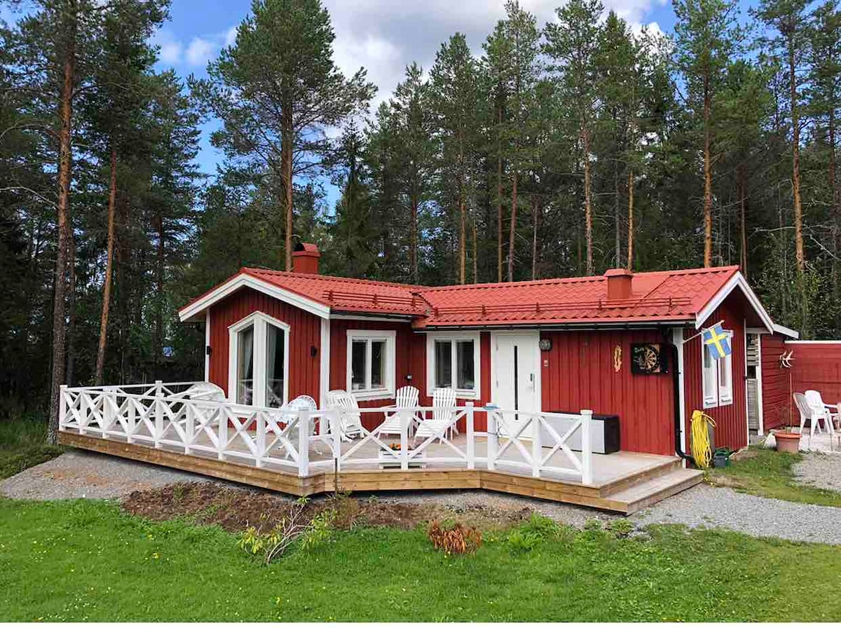 A red wooden cottage is surrounded by trees, featuring a large deck with white chairs and a railing. The welcoming facade includes several windows, allowing natural light to brighten the interior. A small lawn with gravel accents completes the outdoor space.