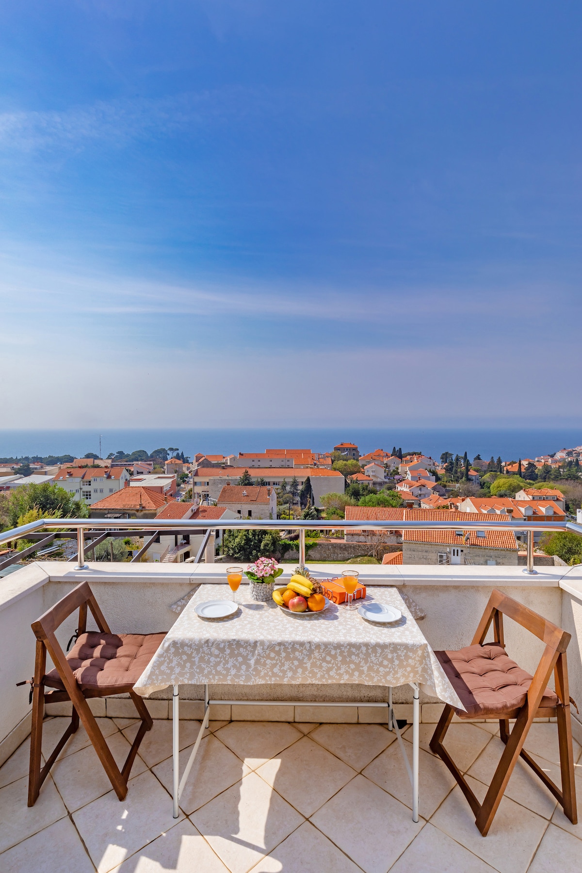 A balcony table is set for two, featuring fresh fruit and drinks. Two wooden chairs surround the table. The scene offers a panoramic view of the surrounding city and sea, under a clear blue sky, showcasing the vibrant atmosphere of the area.