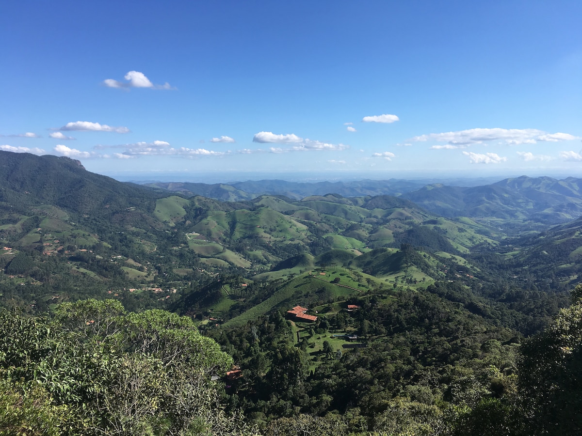 A panoramic view of lush green mountains and valleys is displayed, with varying shades of green. Blue skies with scattered clouds provide a serene backdrop, enhancing the natural landscape's beauty. The distant hills and valleys create depth, showcasing the tranquil setting of the surrounding area.