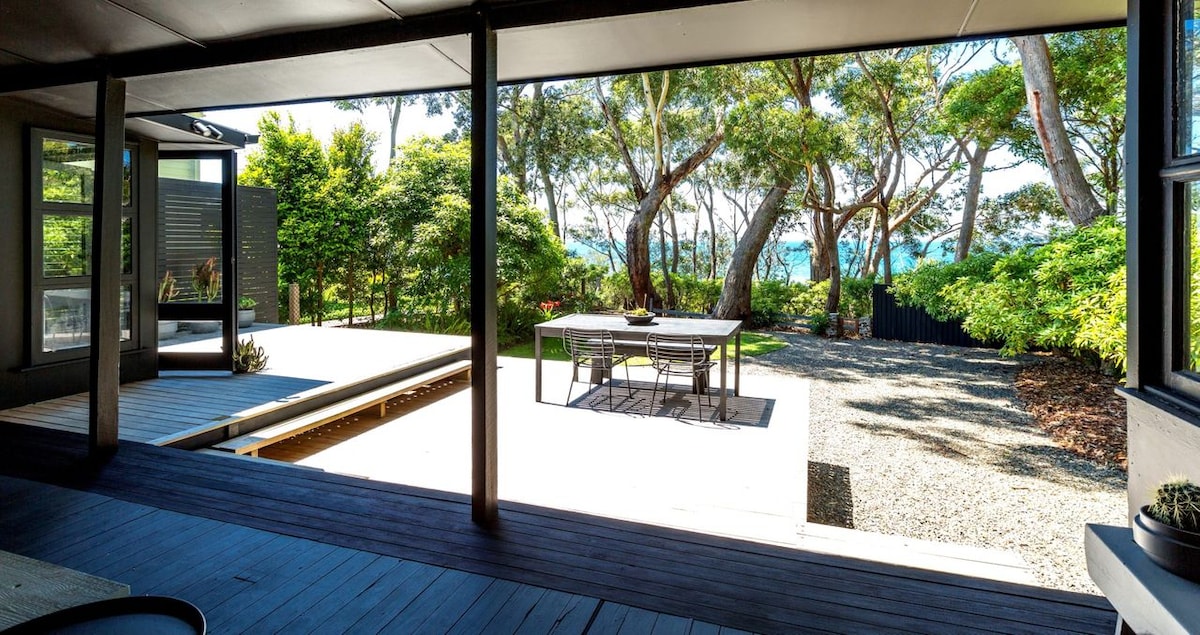 A spacious outdoor area features a wooden deck with a dining table set for four and surrounded by lush greenery. The view reveals a clear glimpse of Jervis Bay through the trees, providing a serene and tranquil setting.